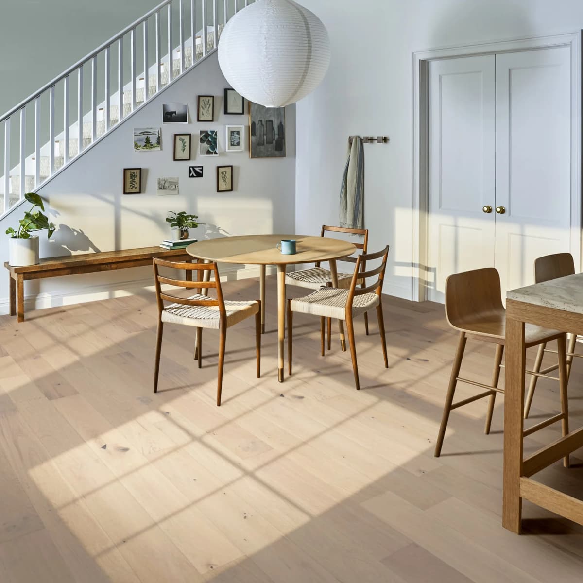 Light white oak engineered hardwood flooring in a sunlit dining and kitchen area with a round table, woven-seat chairs, and a staircase