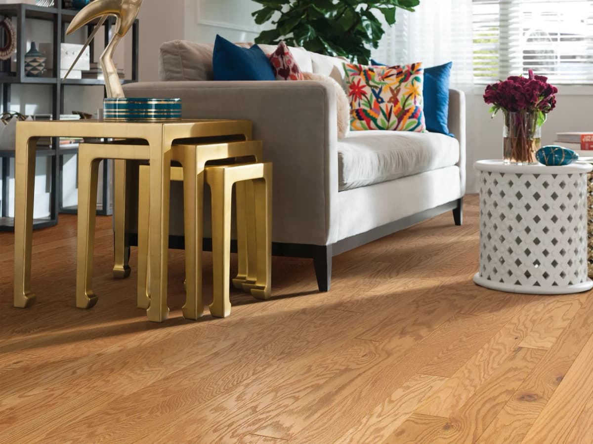 Narrow-plank caramel oak engineered hardwood flooring in a sunlit living room with a cream sofa and gold accent tables