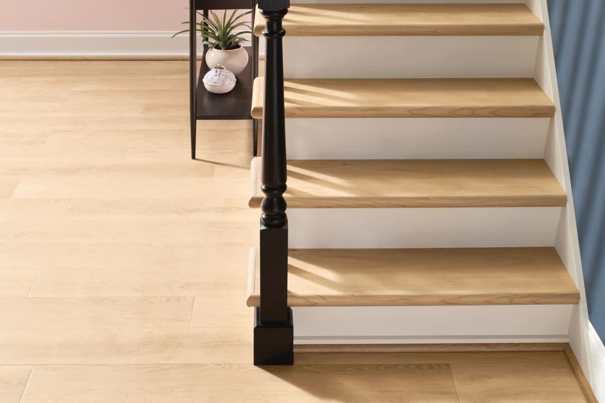 COREtec Original Premium luxury vinyl plank in Wheat Oak at the foot of a staircase with wood-look treads, white risers, and a dark newel post in streaming natural light