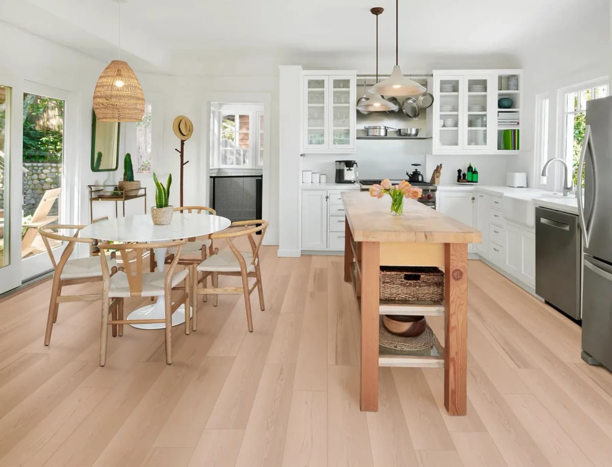 An open kitchen and dining area with light maple-look luxury vinyl plank flooring, a butcher-block island, white cabinetry, a round white table and a woven pendant light