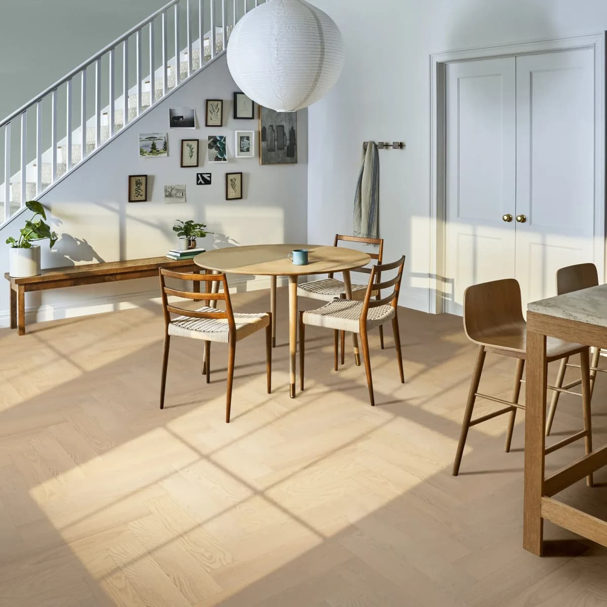 Light European ash engineered hardwood laid in a herringbone pattern under a round wood dining table and woven-seat chairs, with sunlight casting window shadows across the floor