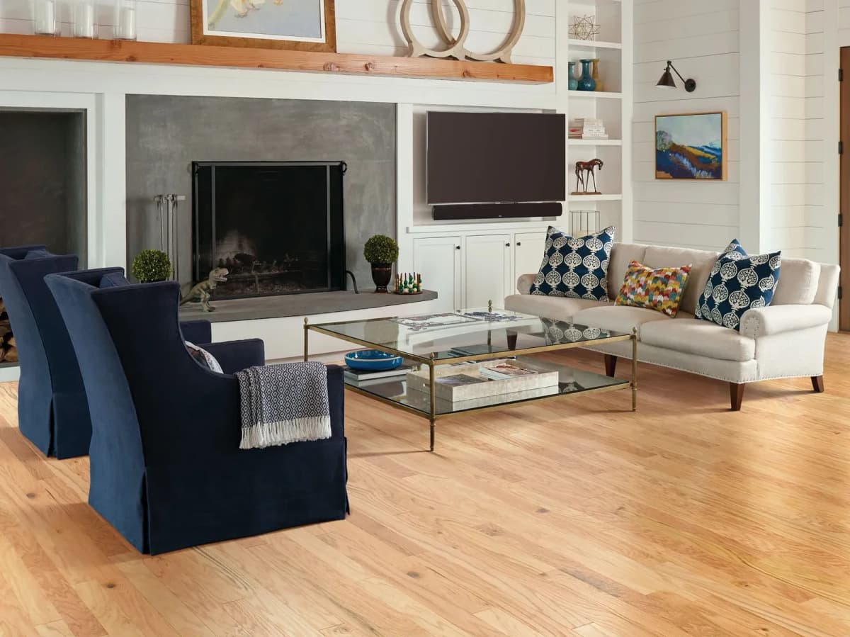 Natural red oak hardwood flooring in a bright living room with two navy armchairs, a cream sofa, a glass coffee table and a fireplace beside built-in shelving