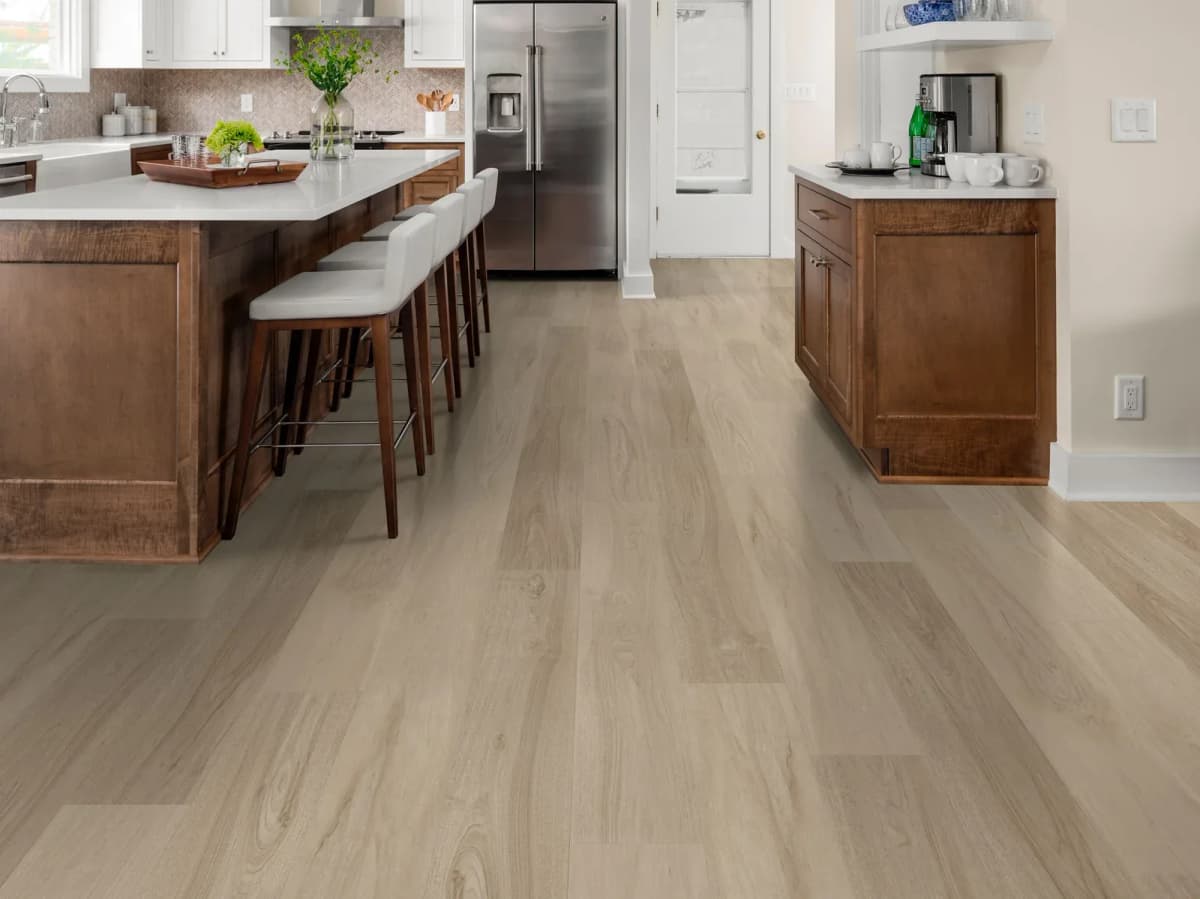 A stainless-steel refrigerator and a wide island with barstools on light luxury vinyl plank flooring in an open kitchen