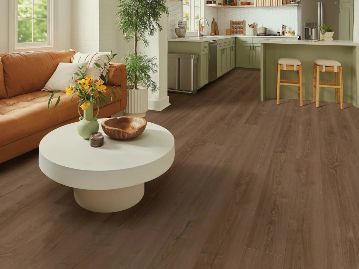 Warm brown luxury vinyl plank flooring running through an open-concept living room and sage-green kitchen, with a cognac leather sofa
