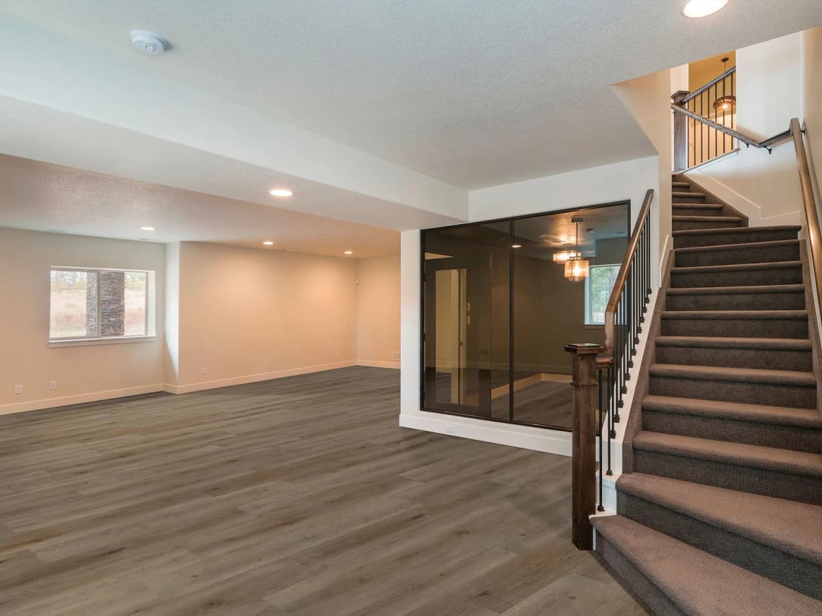 Wood-look luxury vinyl plank flooring covering an open rec room slab, with a staircase and glass-walled room in the background