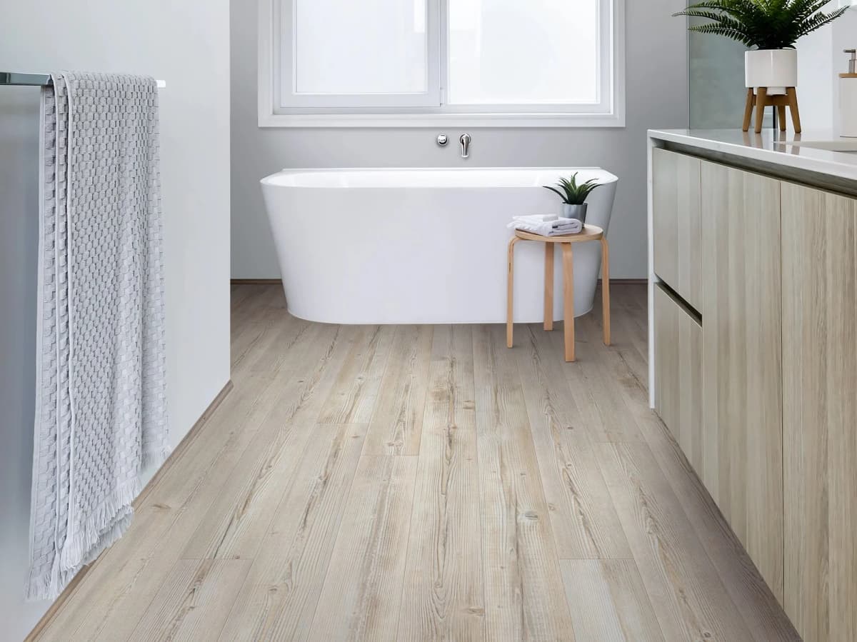 Washed Pine COREtec luxury vinyl plank flooring stretches across a bright bathroom with a freestanding tub, wooden vanity and natural light from a large window