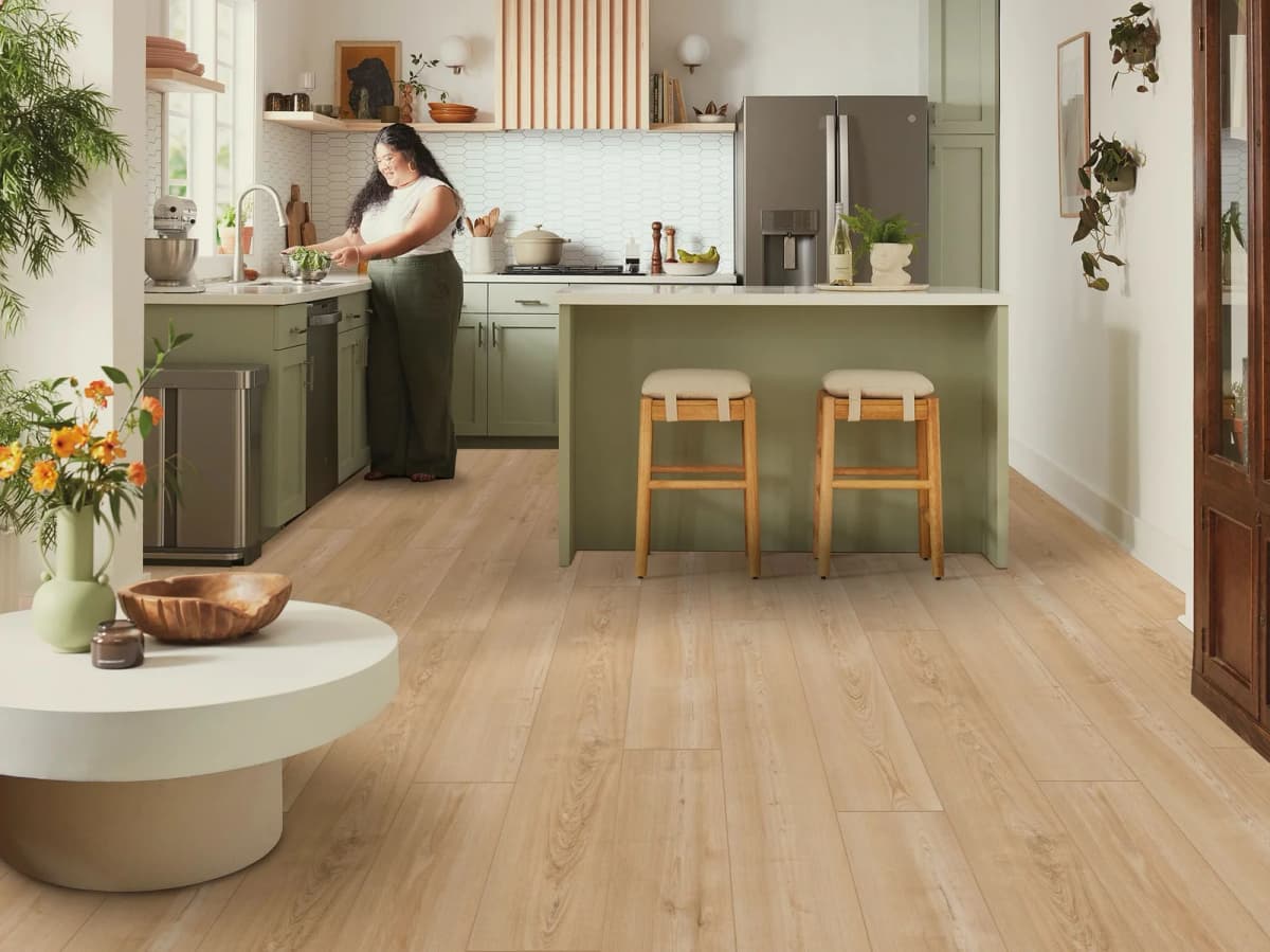 A bright sage-green kitchen with a waterfall island, wooden bar stools, and warm light wood-look luxury vinyl plank flooring underfoot