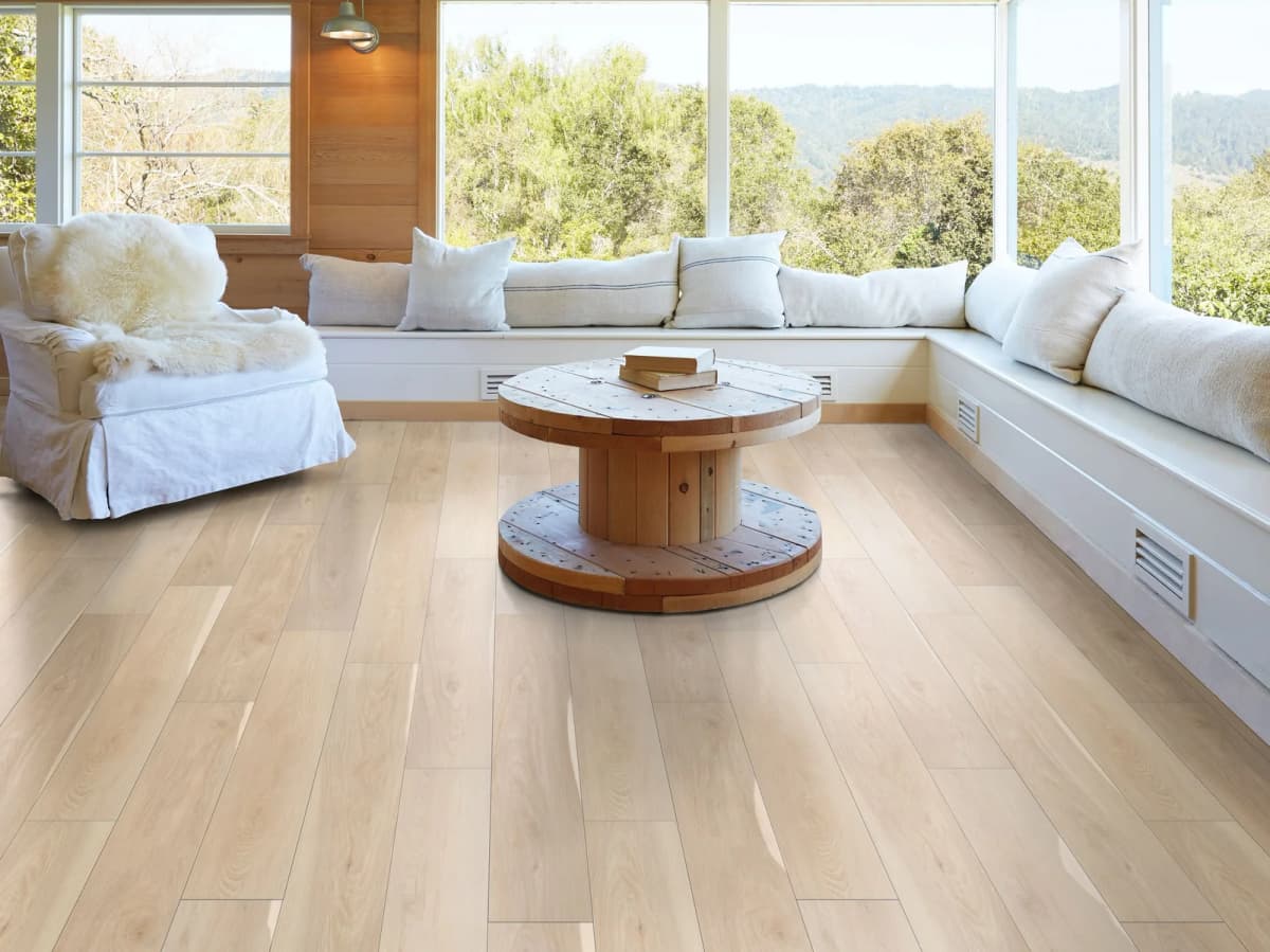 A sunlit great room with floor-to-ceiling windows overlooking trees and mountains, a slipcovered armchair, a built-in window seat, and light oak-look luxury vinyl plank flooring