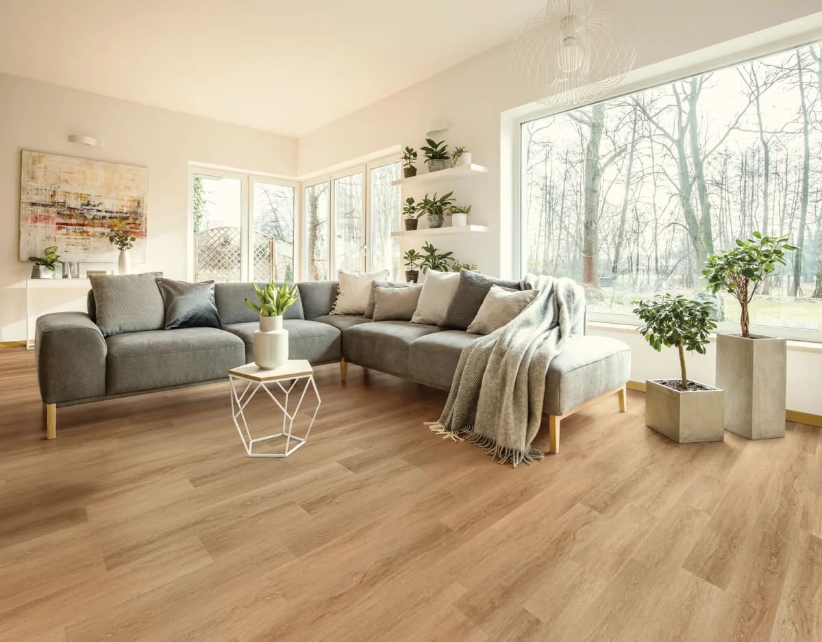 A plant-filled living room with a wall of windows facing bare winter trees, a gray sectional sofa, and warm oak-look luxury vinyl plank flooring