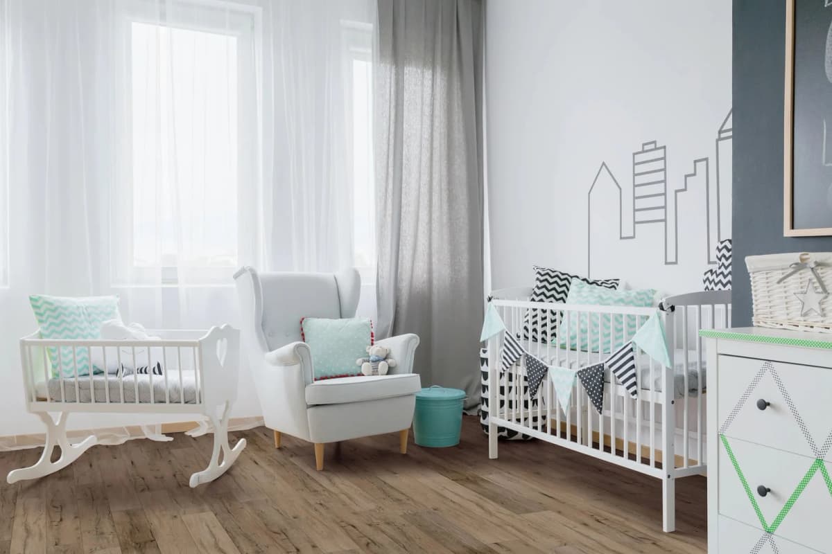 A bright nursery with warm oak-look luxury vinyl plank flooring, two white cribs, a gray wingback chair, and sheer curtains filtering soft natural light