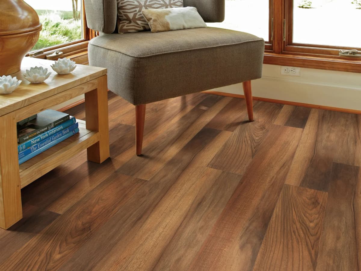 Warm-toned wood-look luxury vinyl plank flooring in a bright sunroom corner with two walls of windows, an upholstered chair, and a wooden side table