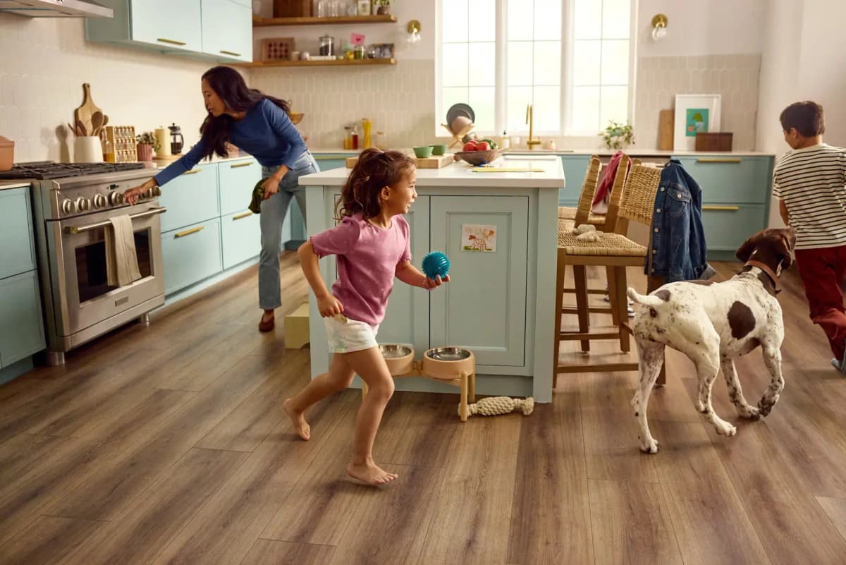 Family kitchen with wide-plank wood flooring — a mother cooks while two kids and their dog play across the floor