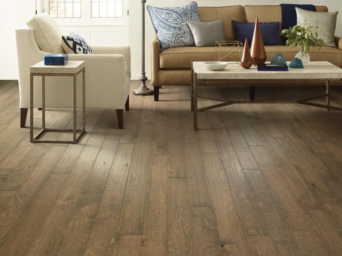 Gray-brown birch plank flooring with a cream armchair and tan leather sofa in a bright living room.