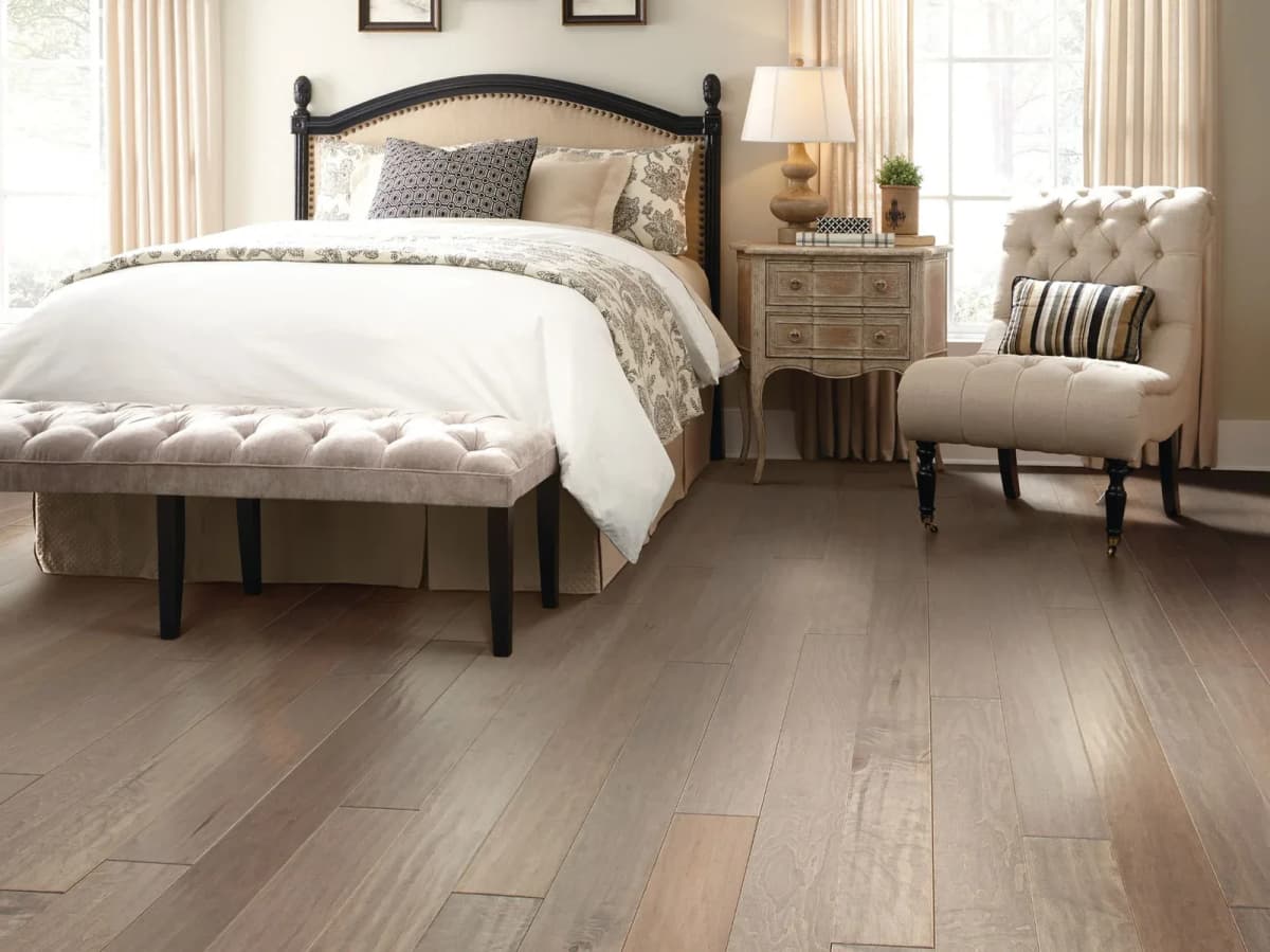 Taupe-brown maple plank flooring with an upholstered bed and bench in a bright bedroom.
