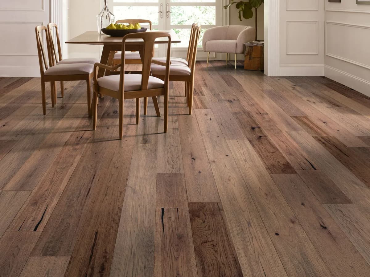 Wide pecan plank flooring in warm mid-brown tones fills a bright dining room with wooden chairs.