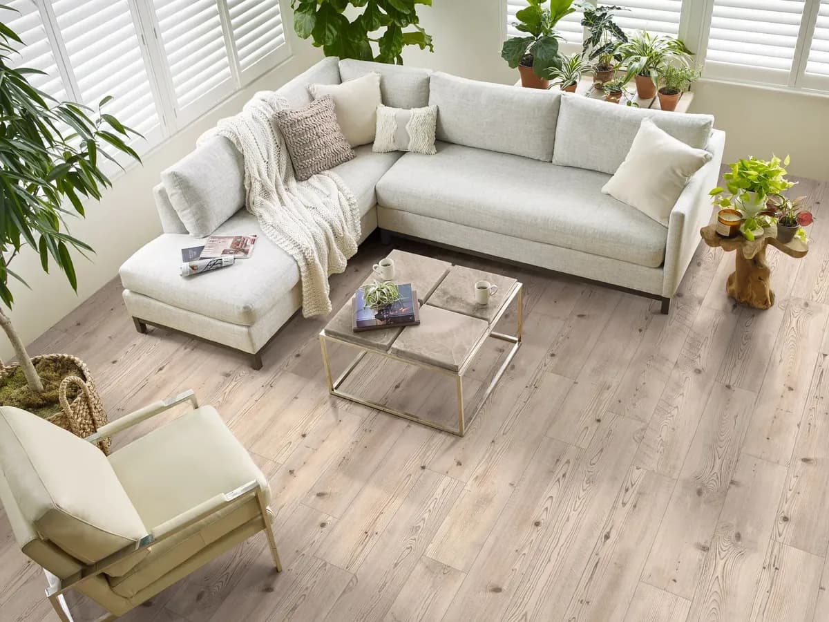 Pale grey-brown luxury vinyl plank flooring fills a bright living room with a cream sectional and potted plants.